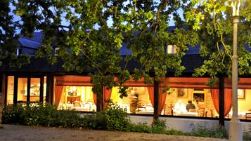 A building with a dining area visible through large windows, surrounded by greenery.