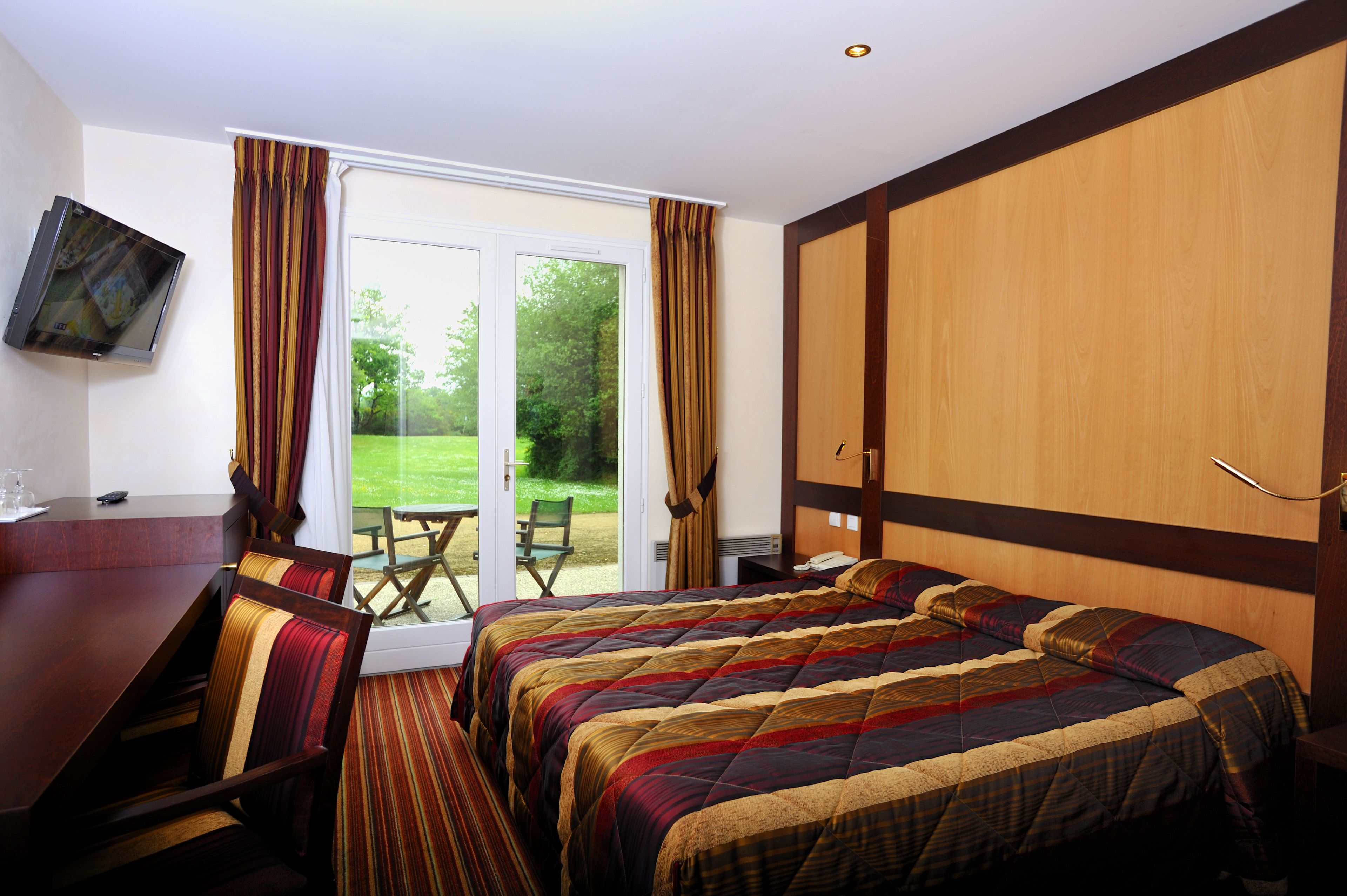 Habitación de hotel con una cama grande, un escritorio y un televisor. Hay una puerta corrediza de vidrio que da a un patio con mesa y sillas.