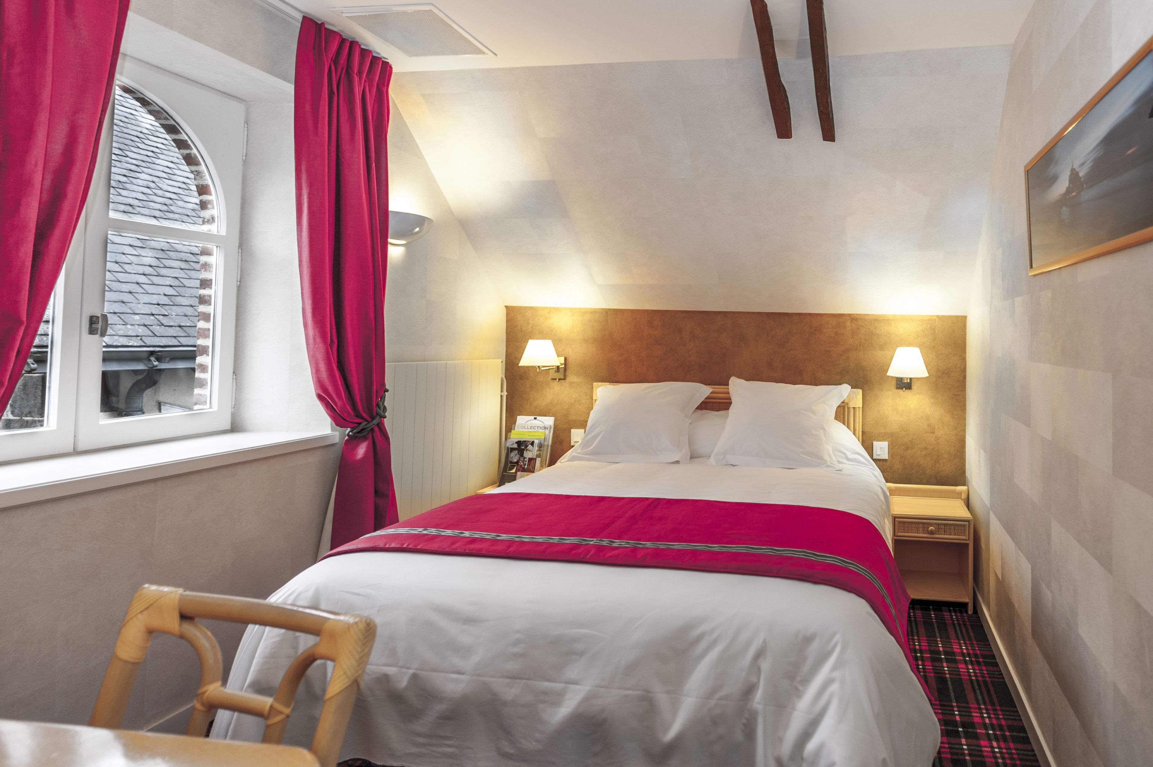A hotel room with a bed, a nightstand, a chair, a window with curtains, and a wall-mounted lamp.