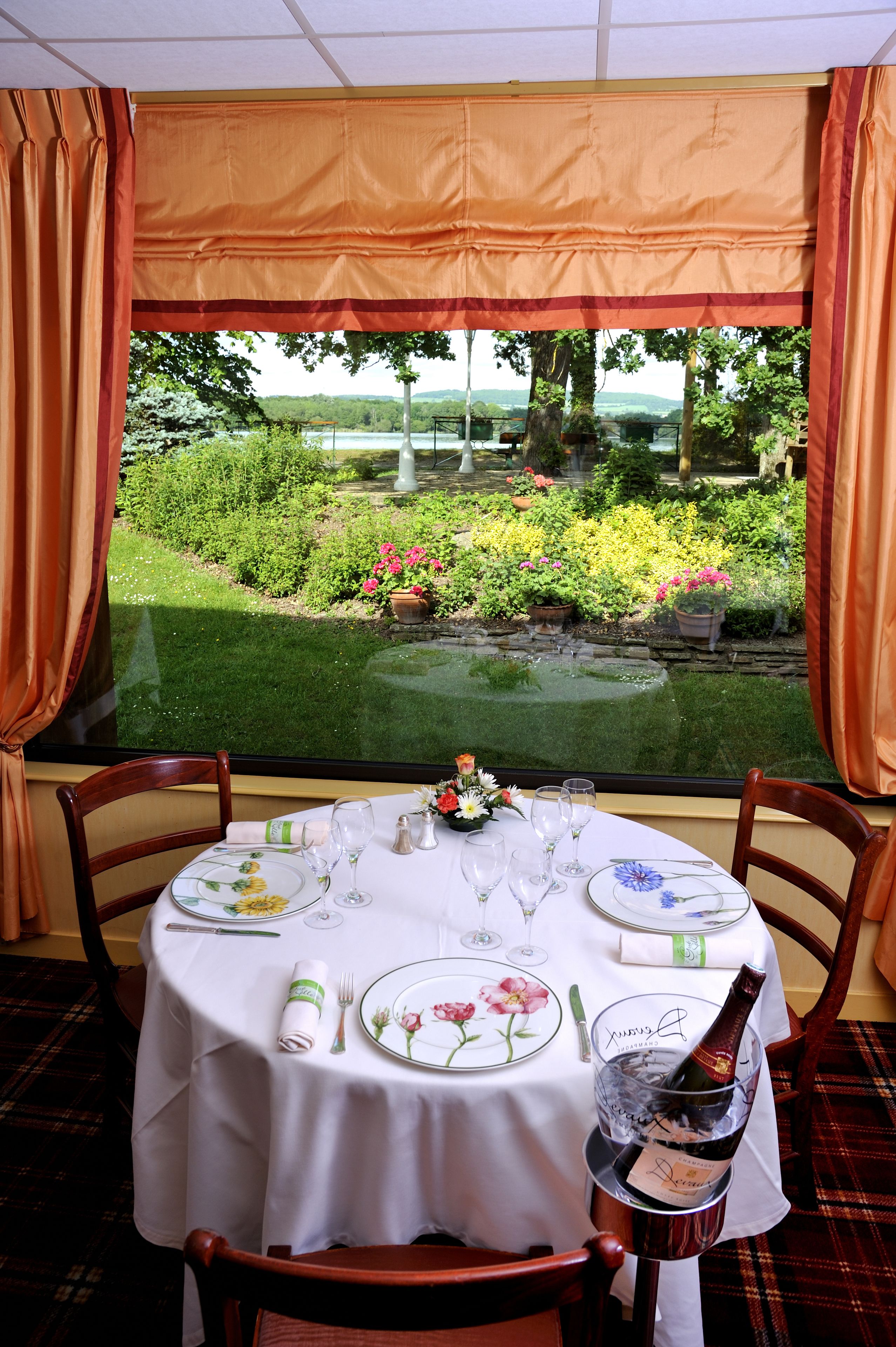 A dining table set for two with a bottle of champagne, a floral centerpiece, and a view of a garden through a window.