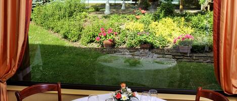 A dining table set for two with a bottle of champagne, a floral centerpiece, and a view of a garden through a window.