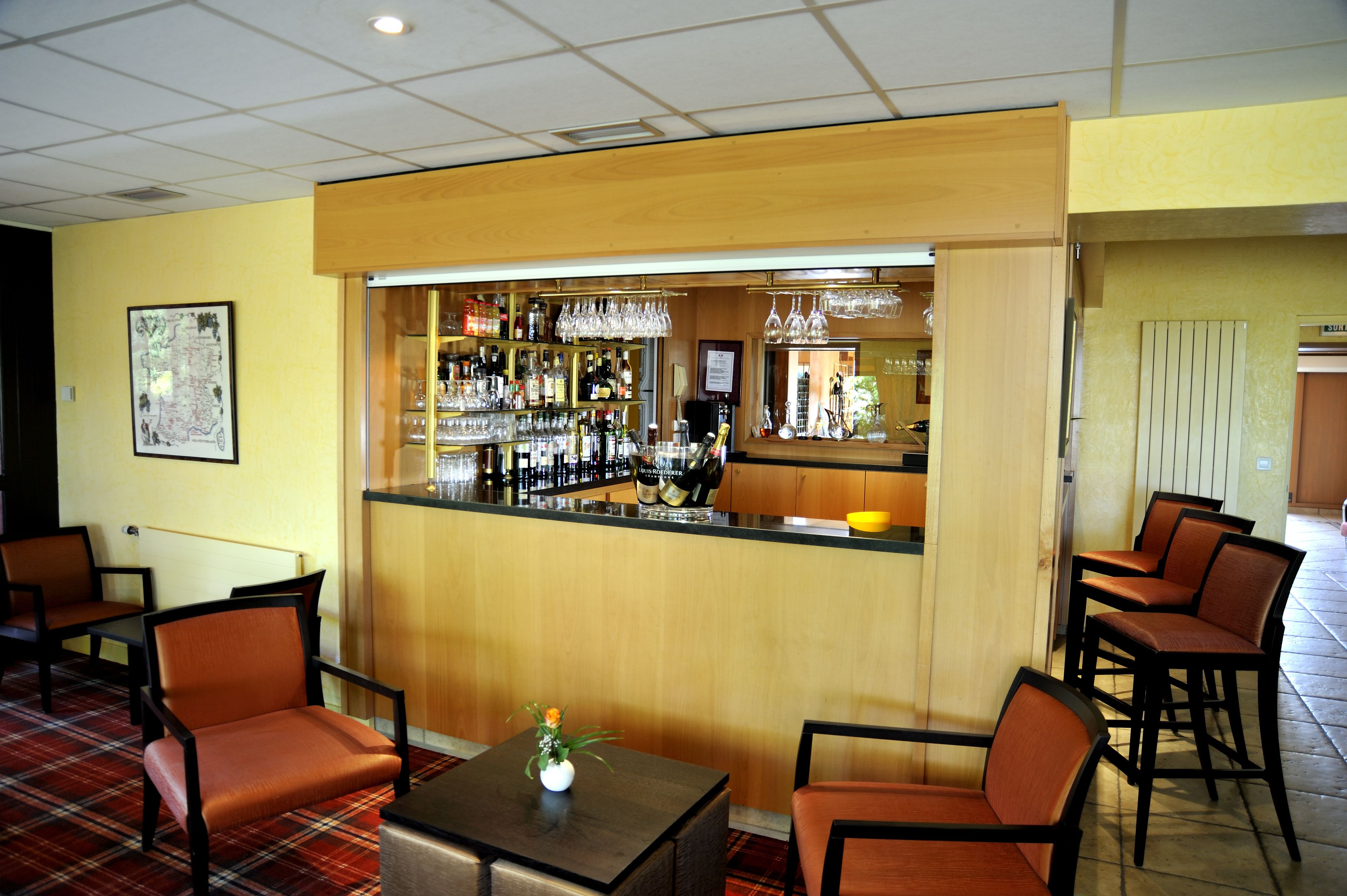 A bar area with a counter, shelves of bottles, glasses, and a small table with a vase. There are chairs around the bar and a map on the wall.