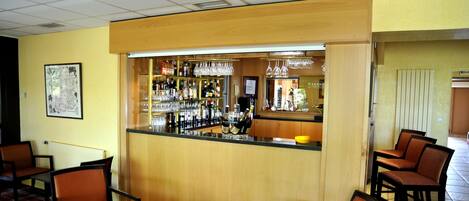 Una zona de bar con mostrador, estanterías llenas de botellas, copas y una mesa pequeña con un florero. Hay sillas alrededor del bar y un mapa en la pared.