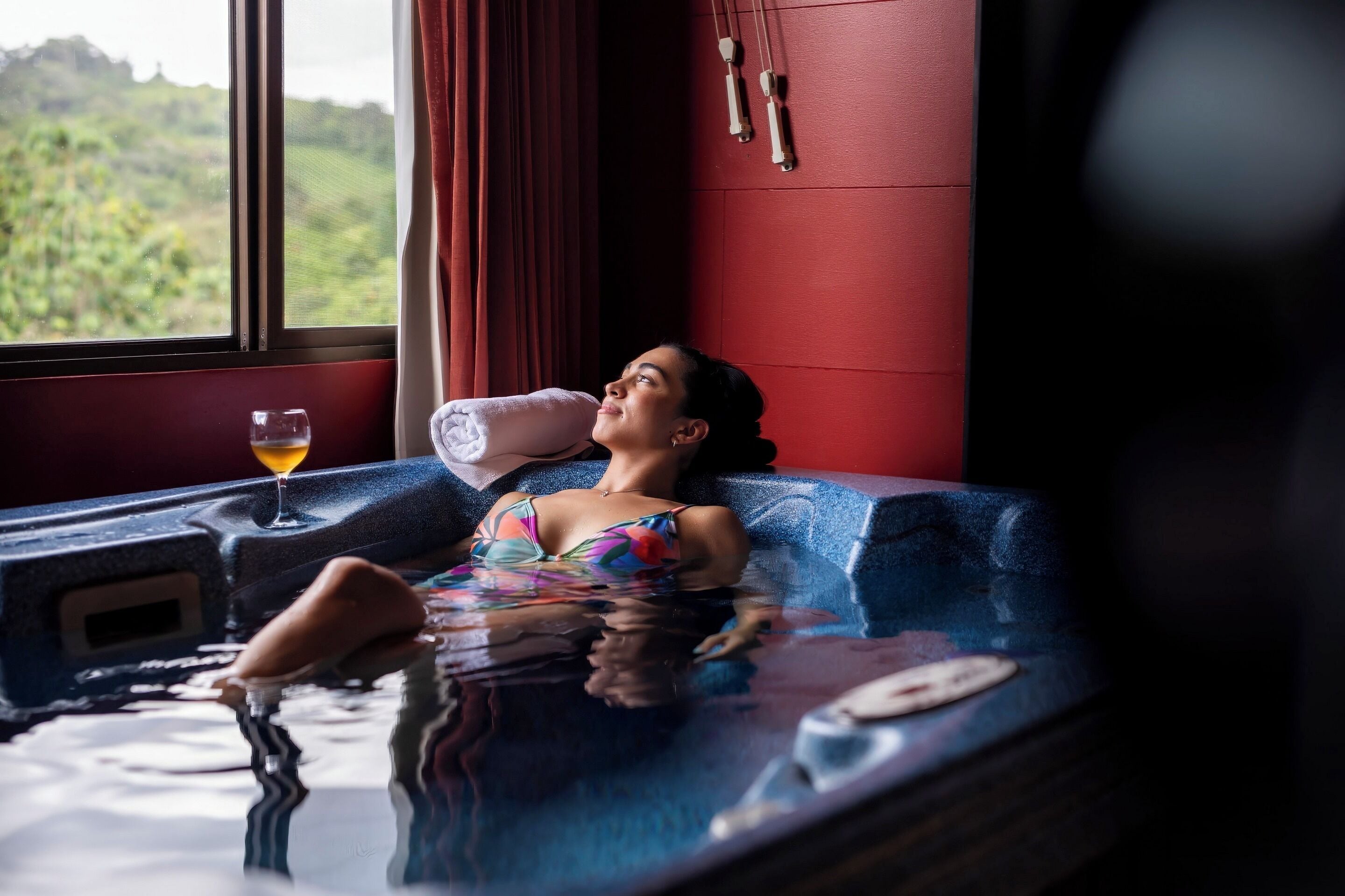 Una persona relajándose en un jacuzzi, una ventana con vistas a la vegetación y una pared roja con un reloj.