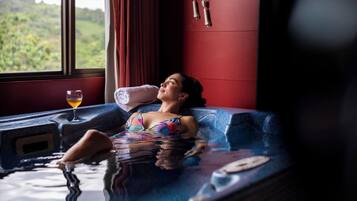 Una persona relajándose en un jacuzzi, una ventana con vistas a la vegetación y una pared roja con un reloj.