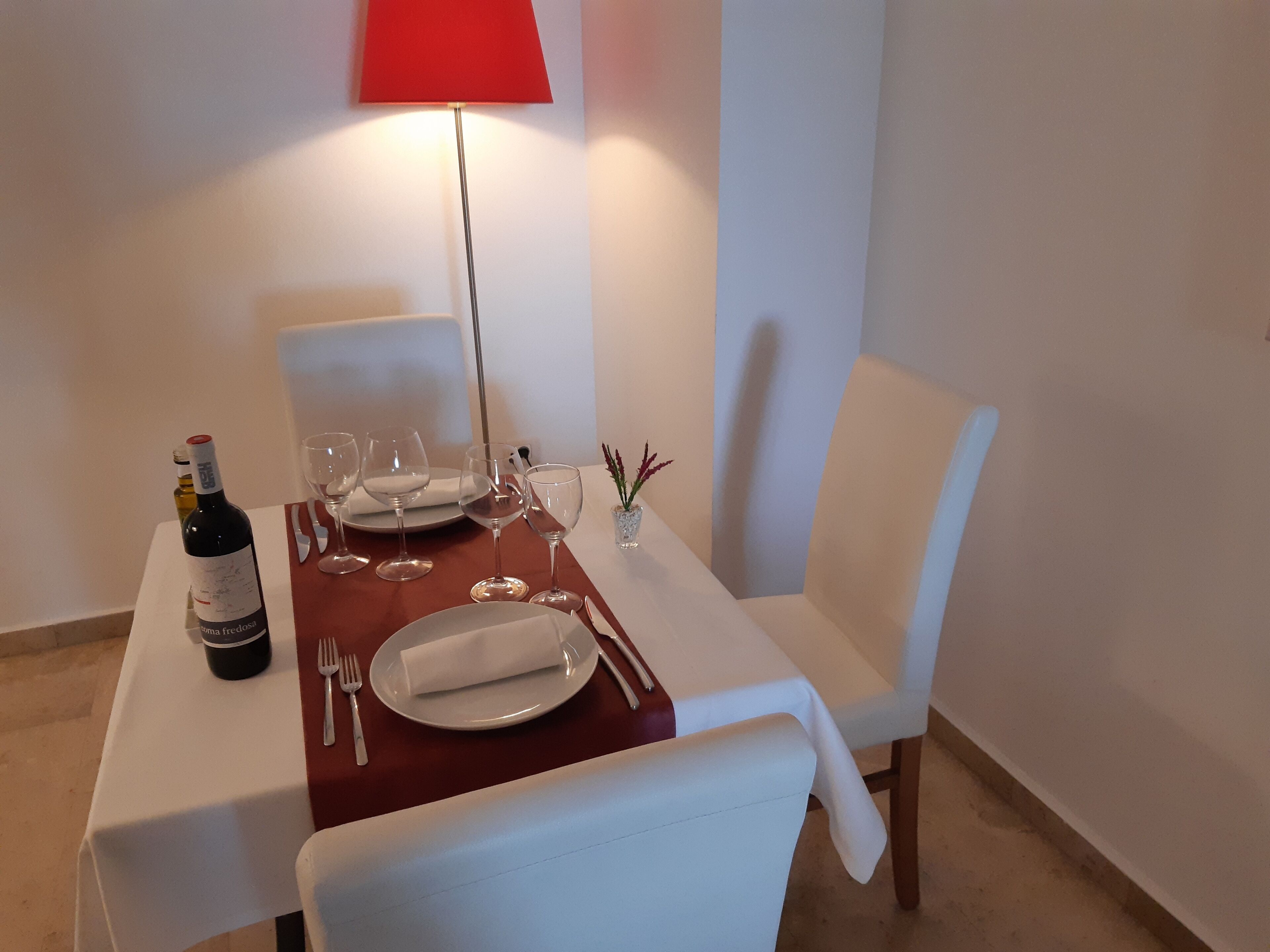 Mesa de comedor dispuesta con mantel blanco, platos, copas de vino y cubiertos. Se ve también una botella de vino y una lámpara roja.