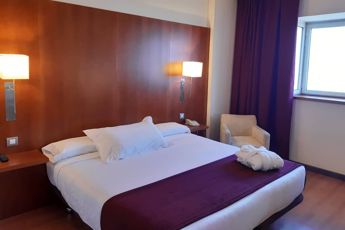 A hotel room with a large bed, two bedside lamps, a chair, a window with curtains, and a nightstand with a phone and remote control.