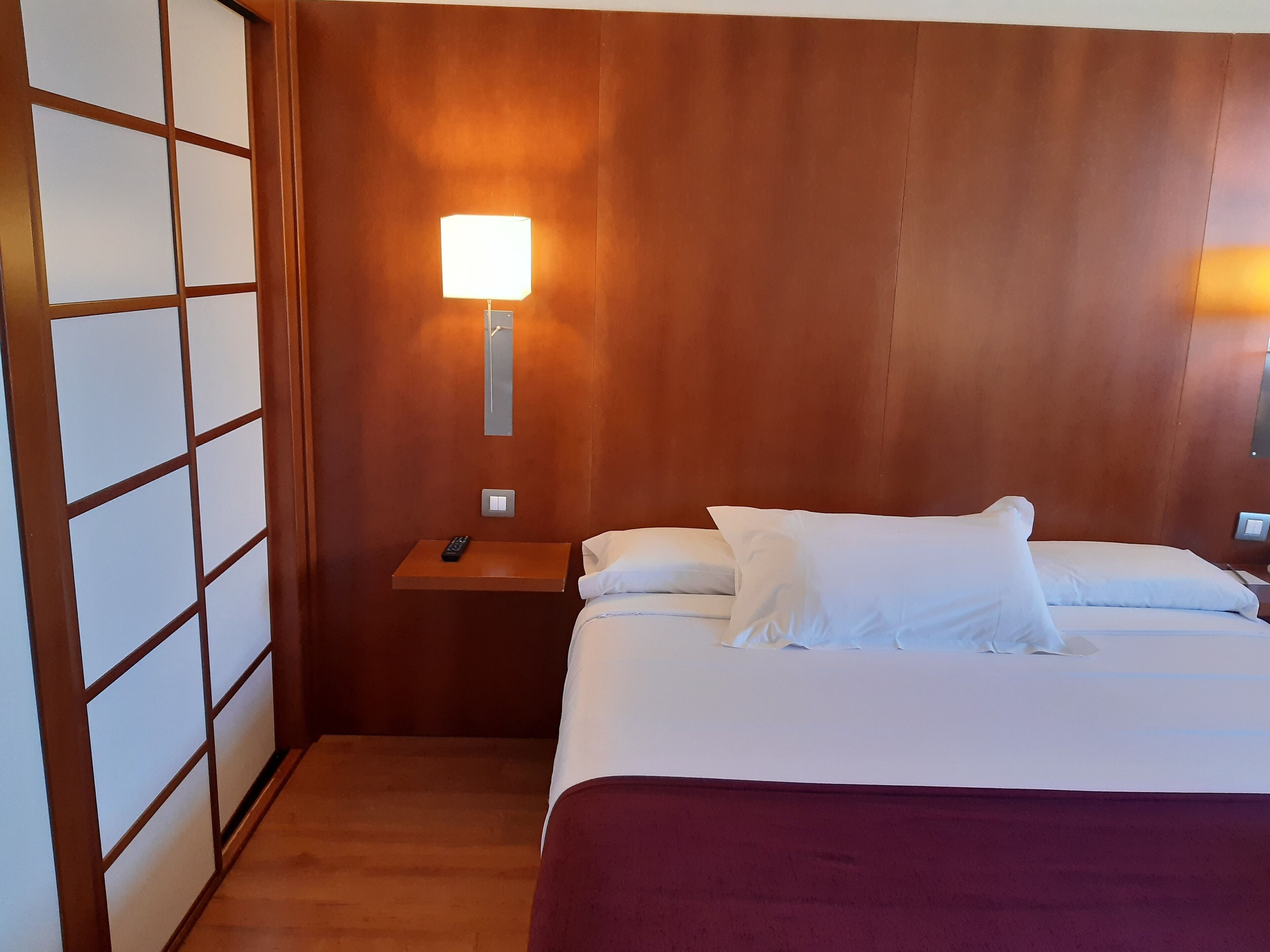 Habitación de hotel con cabecera de madera, cama con ropa blanca, mesita de noche con control remoto y lámpara fijada a la pared.