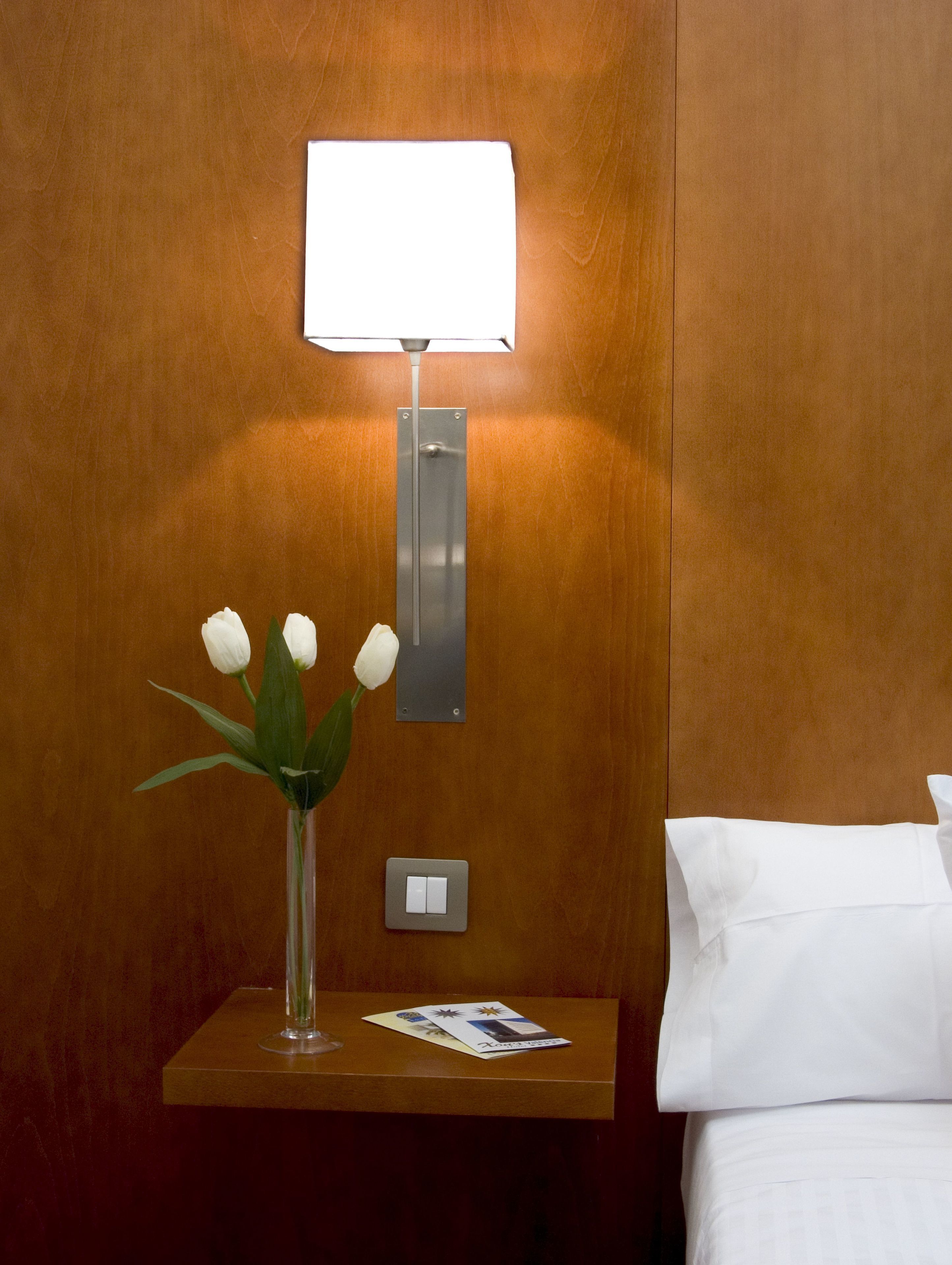 Pared de madera con una mesita de noche, una lámpara, un florero con flores blancas, un interruptor y una revista.