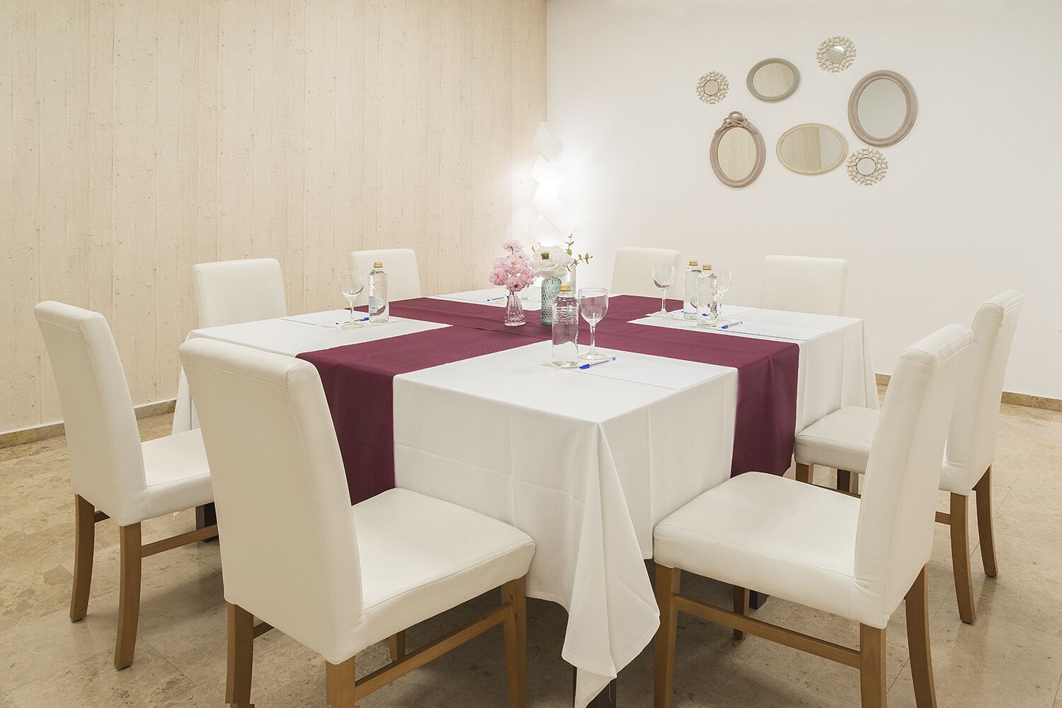 A round table with a white tablecloth and a maroon runner, surrounded by white chairs with wooden legs.