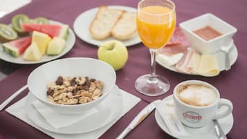 Une table mise pour le petit-déjeuner avec un bol de céréales, un verre de jus d’orange, une tasse de café, ainsi que des assiettes contenant des tartines, des fruits et du fromage.