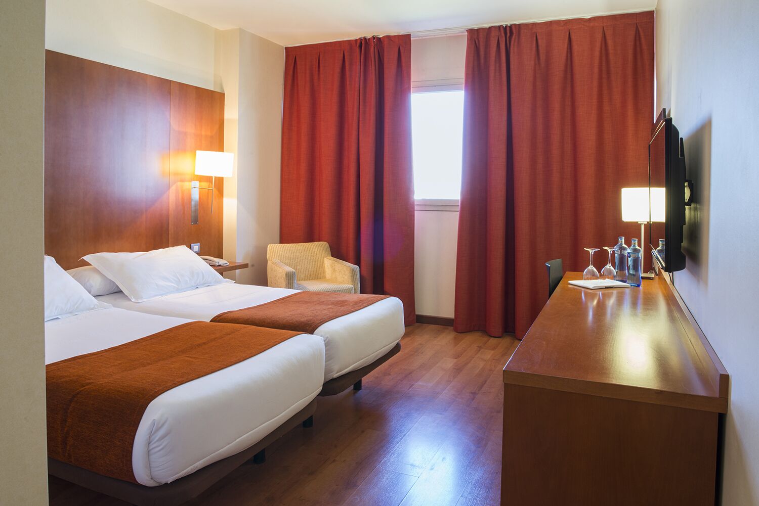 A hotel room with two beds, a desk, a chair, a TV, and a window with red curtains.