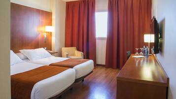 A hotel room with two beds, a desk, a chair, a TV, and a window with red curtains.