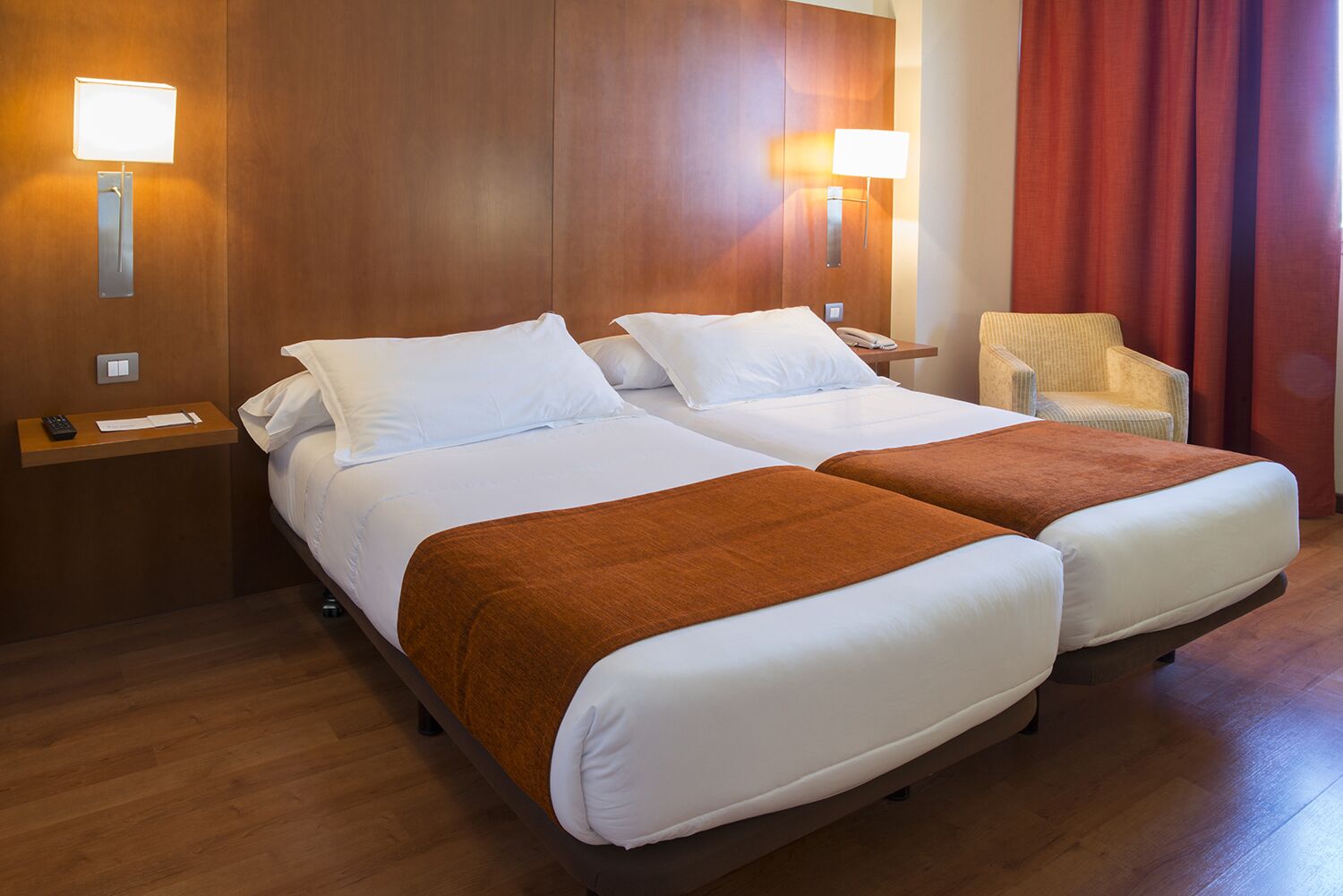 A hotel room with two beds, wooden headboard, bedside tables with lamps, a chair, and a red curtain.