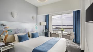 Hotelkamer met een groot bed, een bureau met lamp, een stoel, een klein tafeltje met een plant en een uitzicht op de stad door grote ramen.