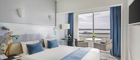 A hotel room with a large bed, a desk with a lamp, a chair, a small table with a plant, and a view of the city through large windows.