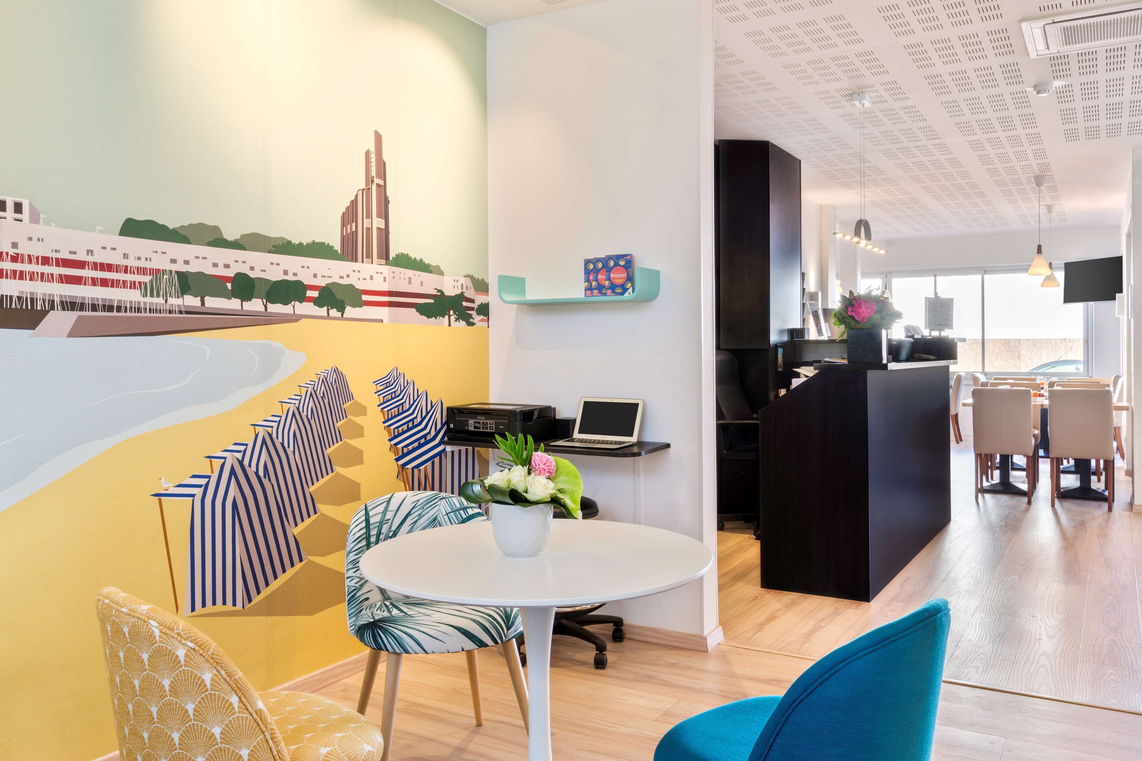A modern hotel lobby with a mural, a reception desk, a round table with chairs, and a dining area in the background.