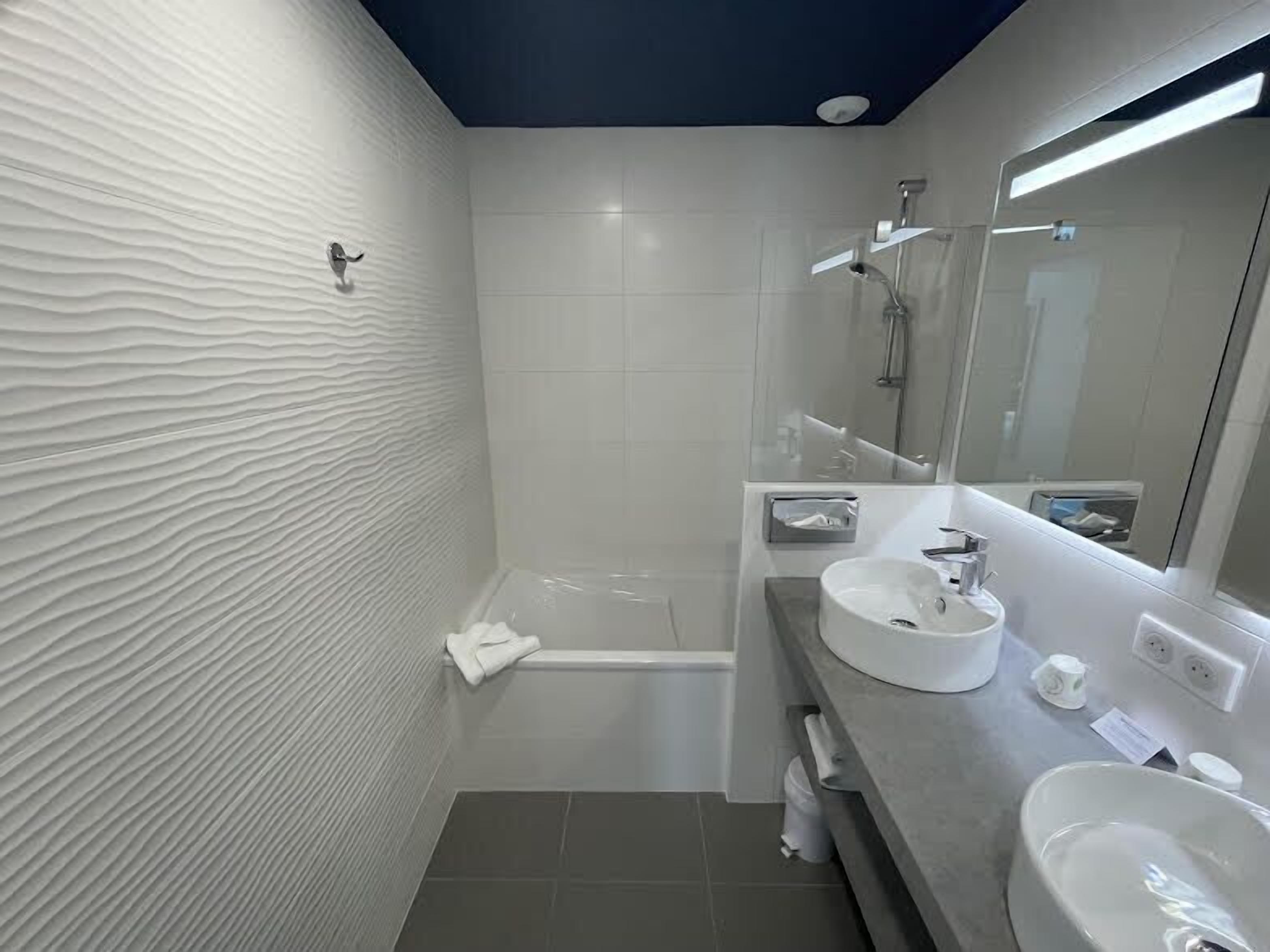 A modern bathroom with a textured wall, a bathtub, a large mirror, and two sinks.