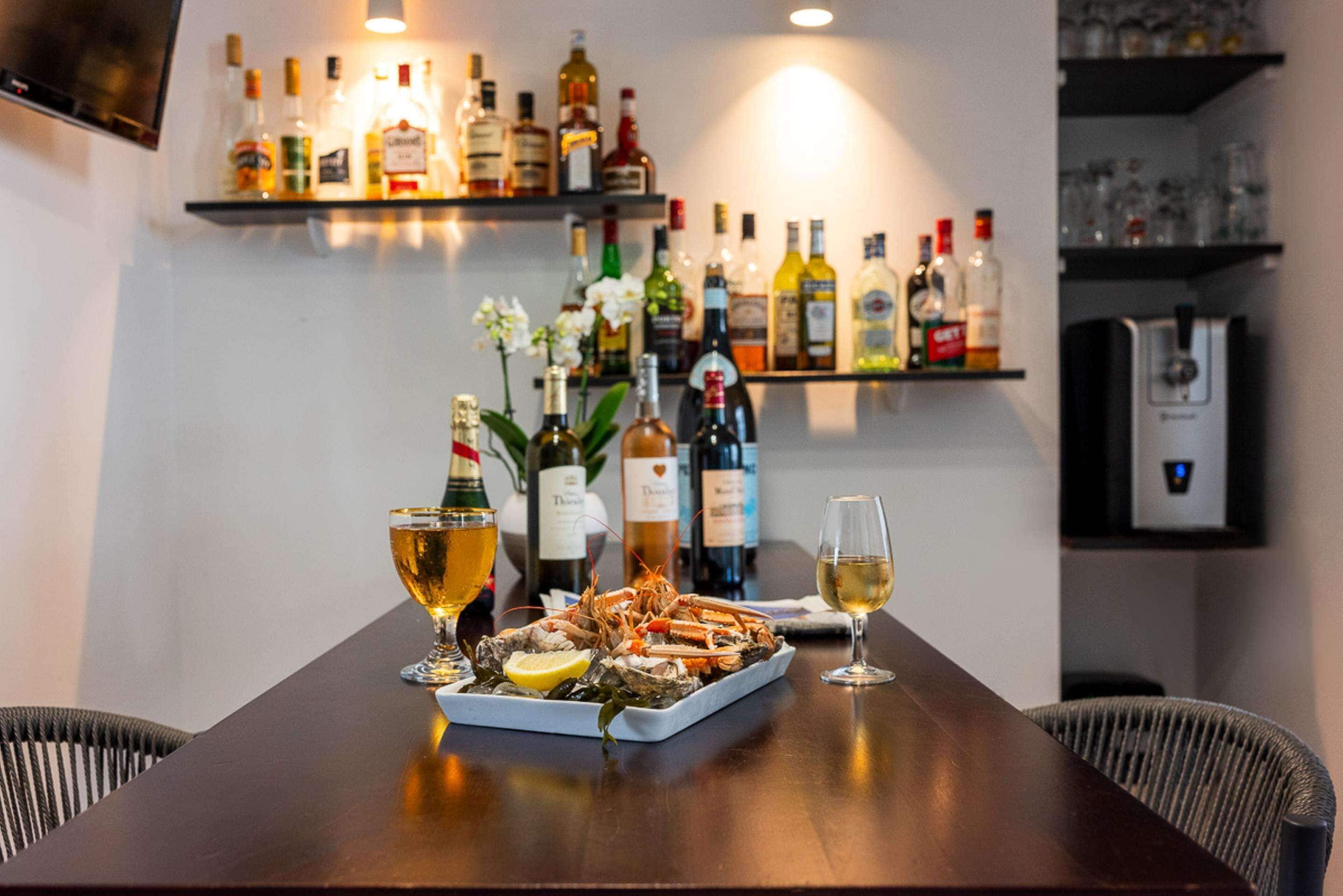 A dining table with seafood, wine glasses, and a variety of liquor bottles on shelves behind.