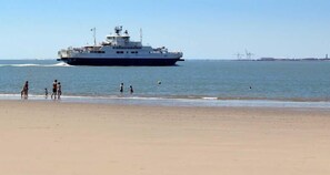 Un grand navire sur l’eau, près d’une plage où des personnes se promènent le long du rivage.