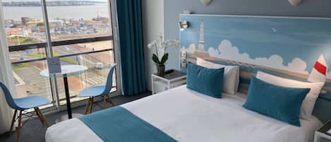 A hotel room with a large bed, two pillows, a nightstand, a chair, a small table, and a view of a cityscape through a large window.