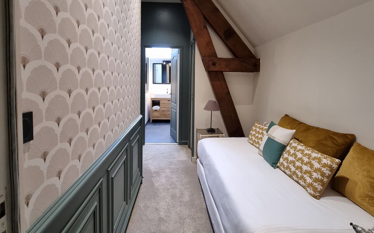 A bedroom with a bed, pillows, a nightstand, and a lamp. There is a hallway leading to another room.