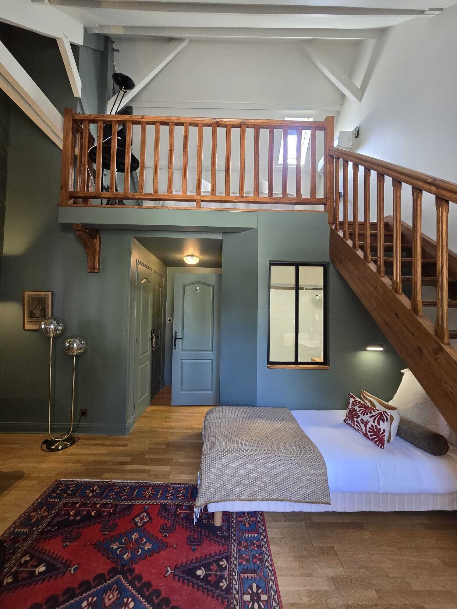 A loft bedroom with a high ceiling, a wooden staircase, a bed with a patterned throw, a red rug, and a floor lamp.