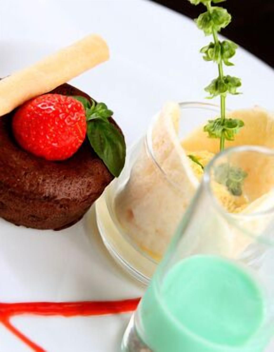 Un plato de postre con pastel de chocolate cubierto con fresa y albahaca, acompañado de un pequeño cuenco de crema y un vaso de una bebida verde.