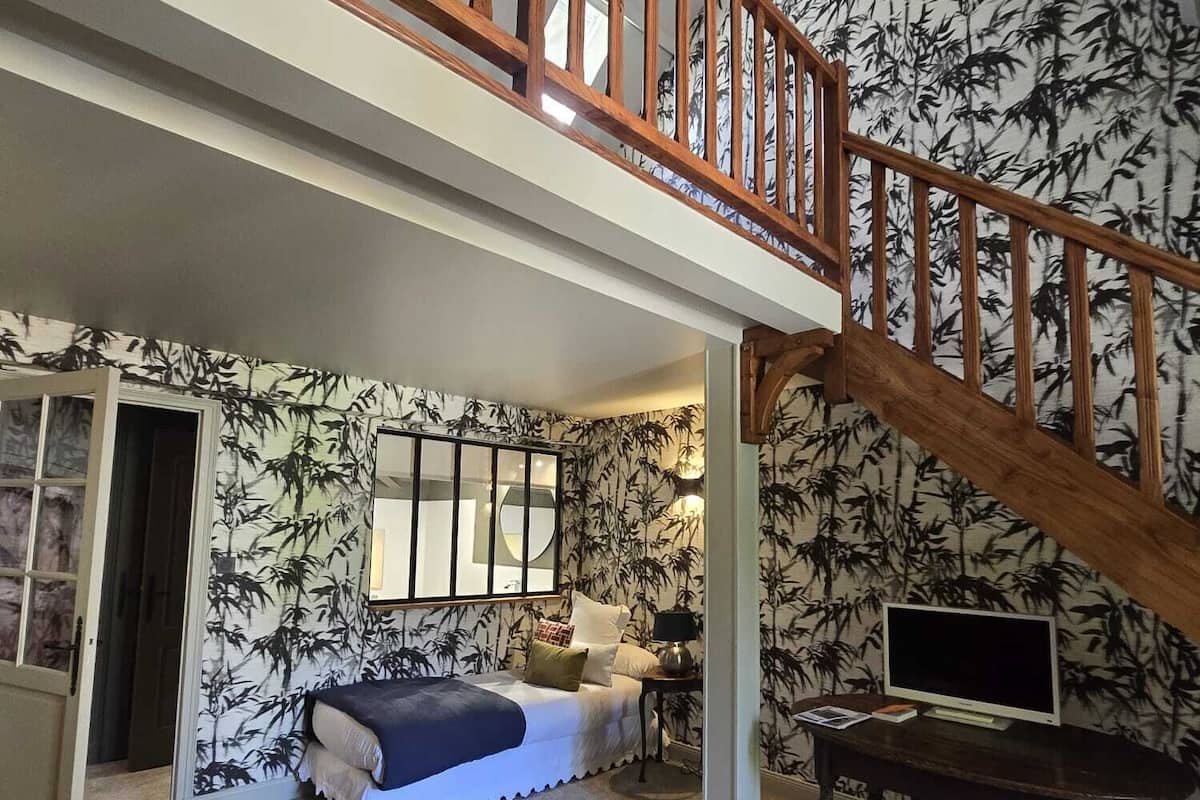 Una habitación amplia con techo alto, una escalera de madera, una cama, un televisor sobre una mesa de madera y un ventanal con vistas.