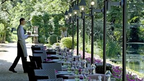 Restaurante com mesas ao ar livre com vista para um lago, mesas montadas com copos e talheres, e um garçom atendendo os clientes.