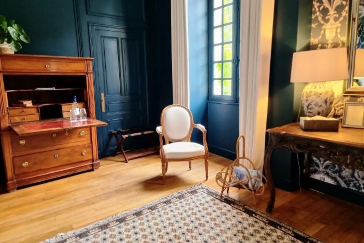 A room with a wooden dresser, a white chair, a patterned rug, and a desk with a lamp.