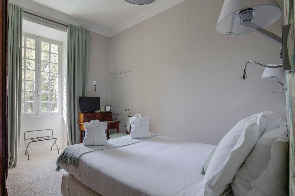 Habitación de hotel con una cama, un escritorio con televisor, una ventana con cortinas y una lámpara fijada a la pared.