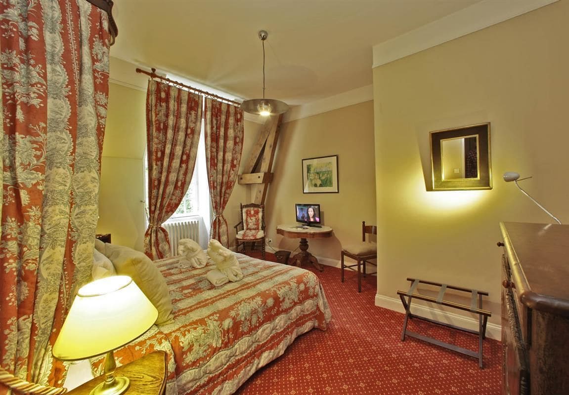 A hotel room with a bed, a desk, a chair, a lamp, a picture frame, and a radiator.