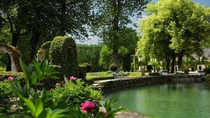 Um jardim tranquilo com um lago, vegetação exuberante e um edifício clássico ao fundo.