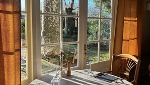 A dining area with a table set for two, large windows, and curtains. - La Sommellerie (Chateauneuf-du-Pape)