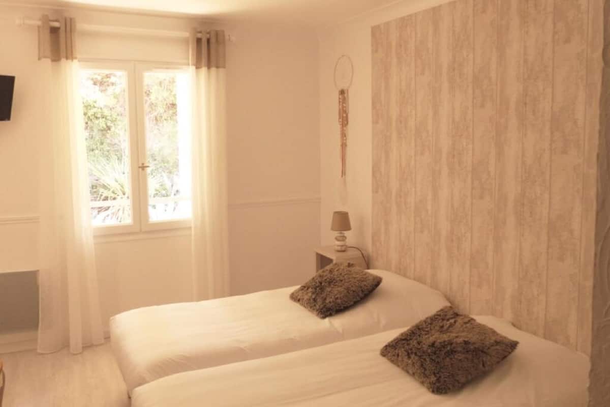 A bedroom with two beds, a window with curtains, a bedside table with a lamp, and a wall-mounted TV.