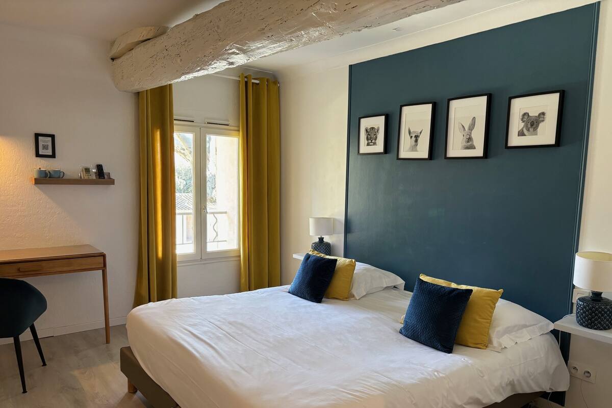 A bedroom with a bed, a desk, a chair, and a window with curtains. There are framed pictures on the wall.