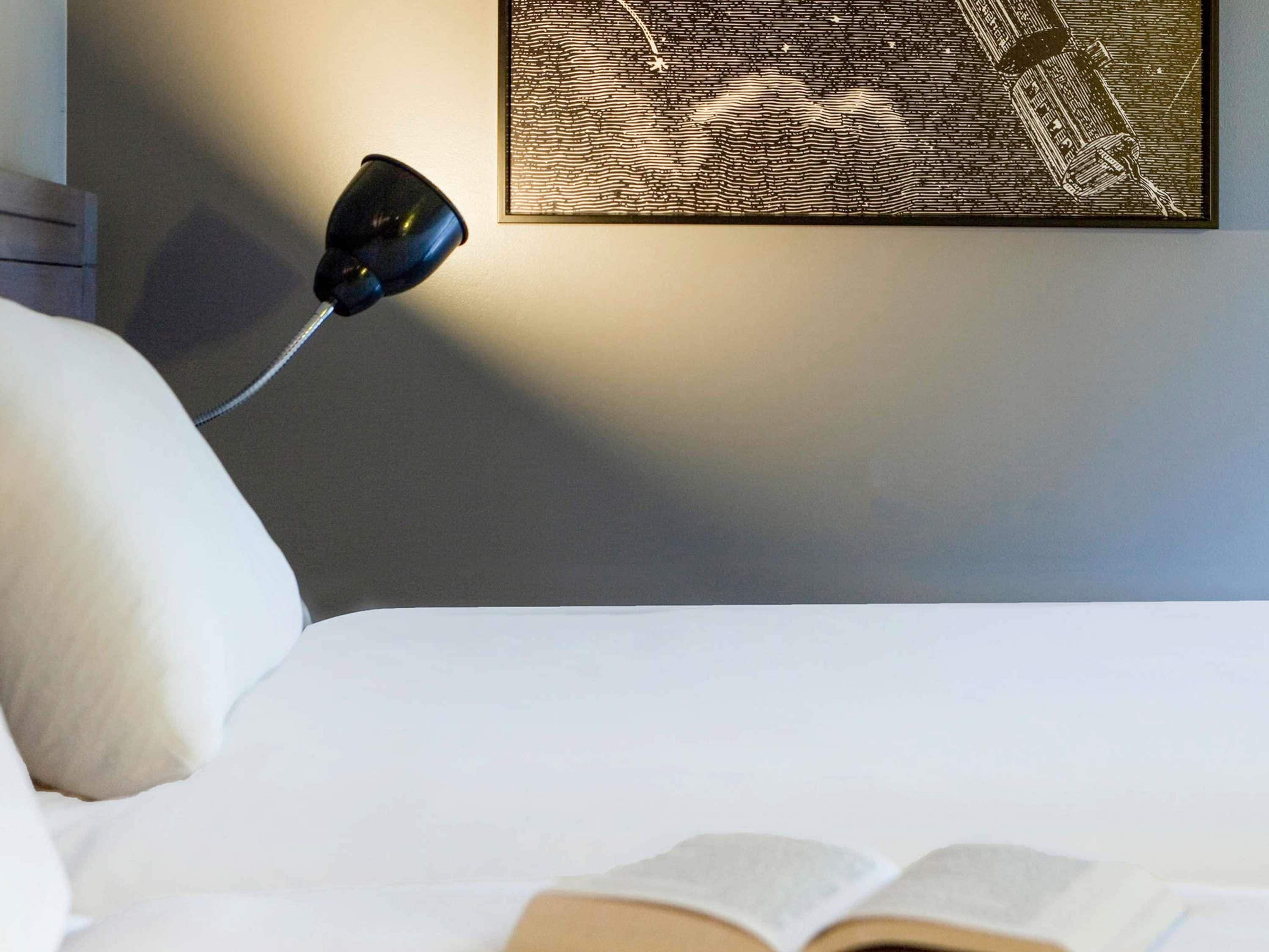 A neatly made bed with a book open on it, a black desk lamp, and a framed artwork on the wall.