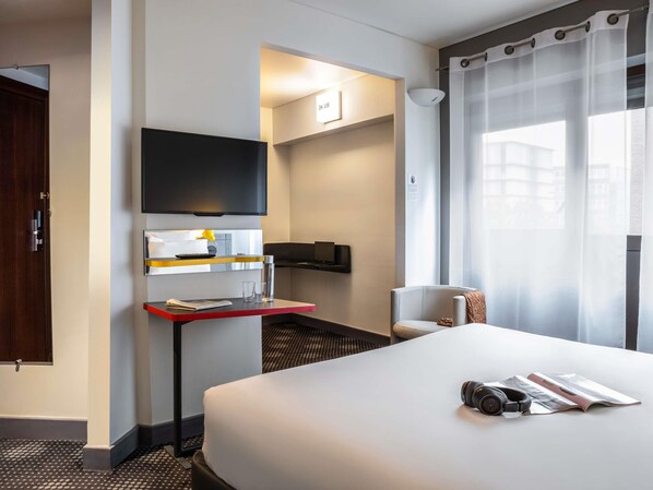 A hotel room with a bed, a desk with a flat-screen TV, a chair, and a window with sheer curtains.
