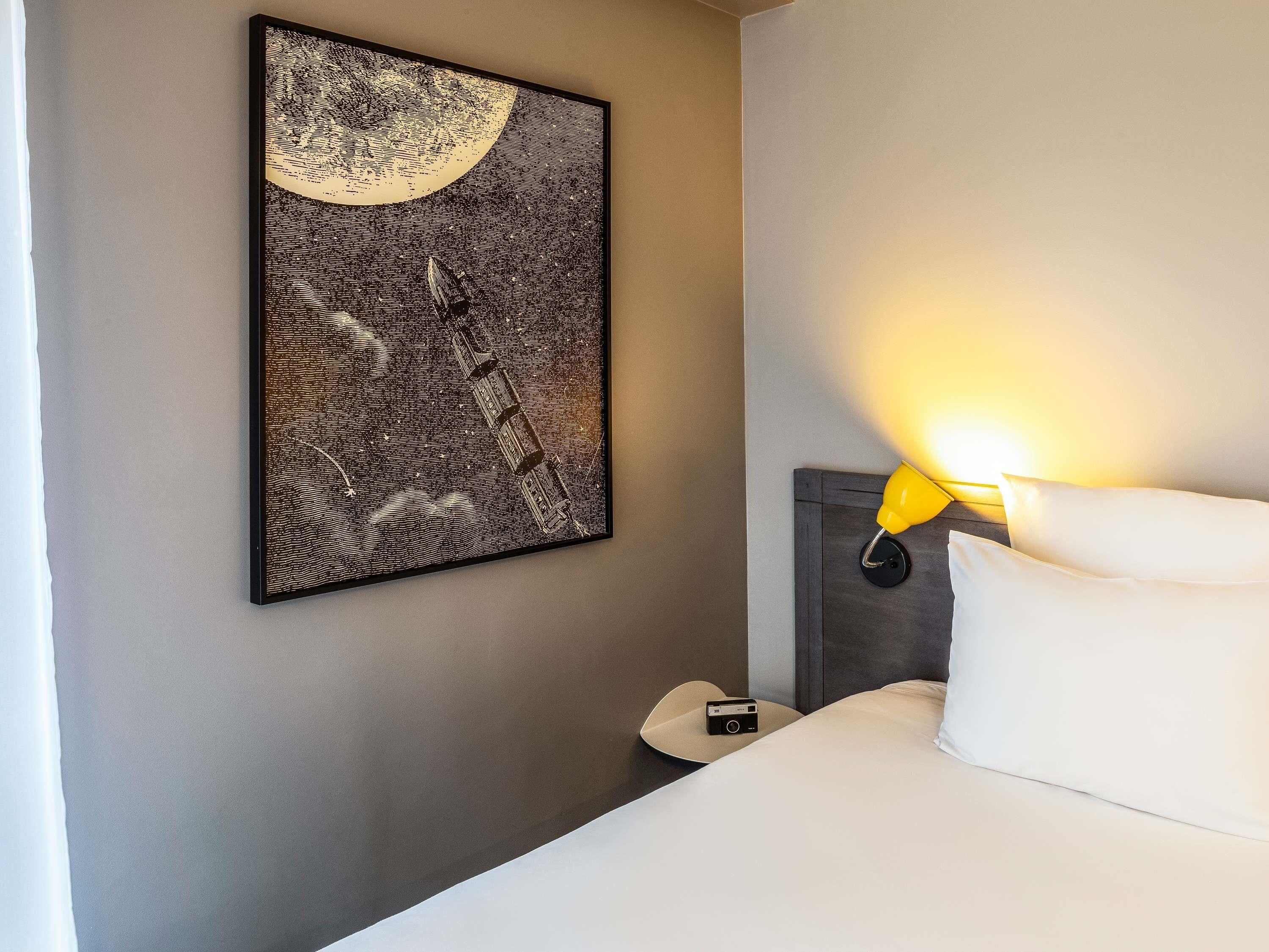 A hotel room with a bed, a nightstand, a lamp, a wall-mounted artwork, and a camera on the floor.