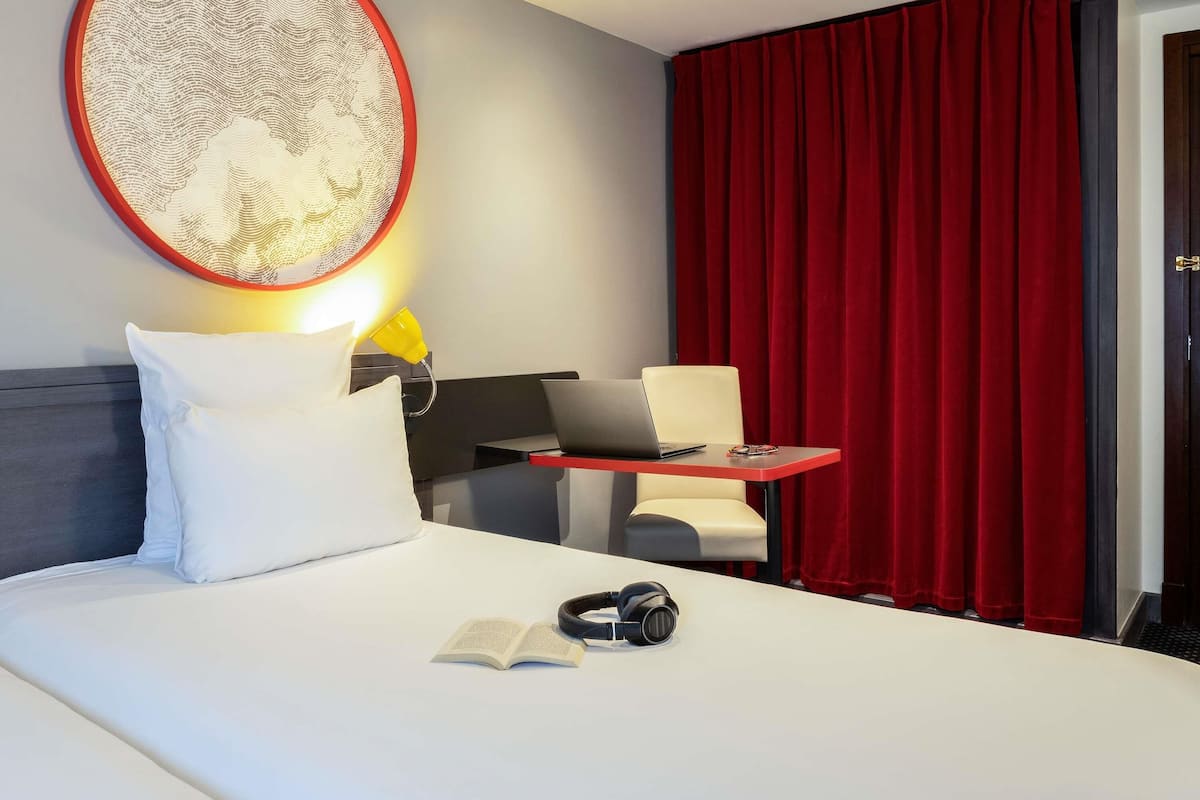 A hotel room with a bed, a desk with a laptop, a chair, a red curtain, and a wall-mounted lamp.