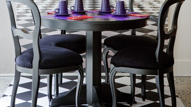 Un coin repas avec un sol à carreaux, une table ronde entourée de chaises, et une vue sur un couloir décoré avec un plafond à carreaux.
