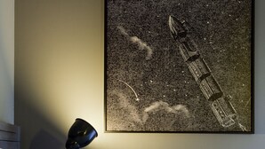 Ein Schlafzimmer mit einem Bett, einem offenen Buch, einem an der Wand befestigten Kunstwerk mit einer Rakete und einem Mond sowie einer Schreibtischlampe.