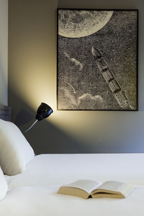 A bedroom with a bed, an open book, a wall-mounted artwork depicting a rocket and moon, and a desk lamp. - ibis Styles Paris Mairie de Montreuil (Montreuil)