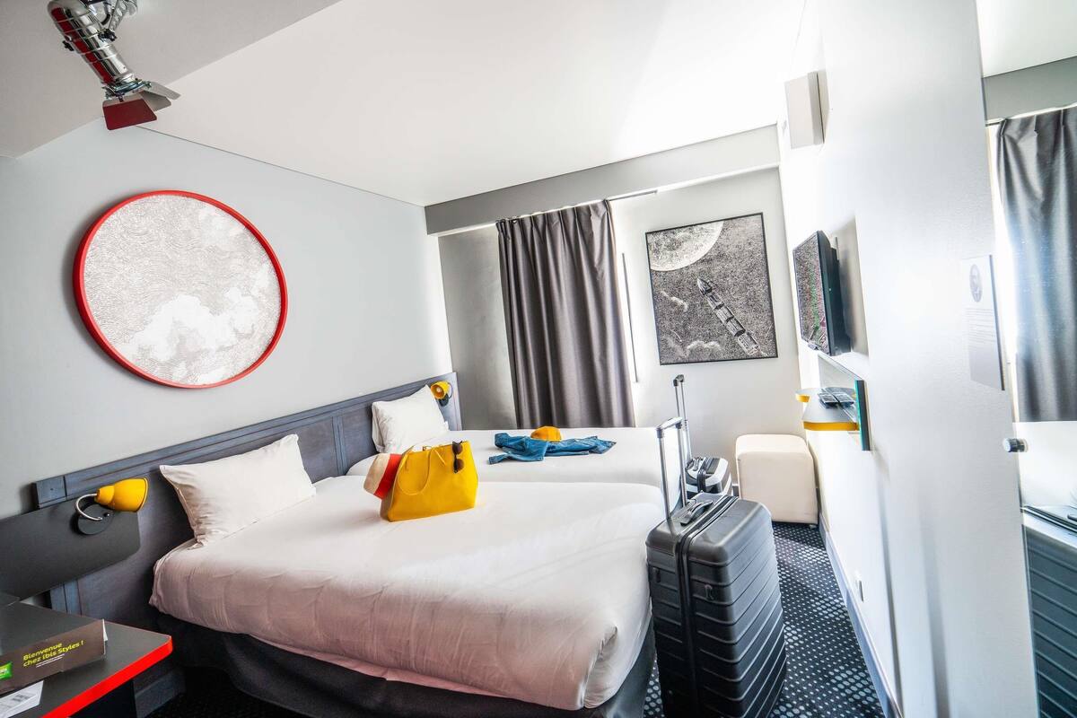 A hotel room with a bed, a desk, a chair, a suitcase, and a window with curtains.