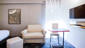 Ein Hotelzimmer mit einem weißen Sofa, einem kleinen Tisch, einem Fernseher, einem Gemälde und einem Fenster mit Vorhängen.