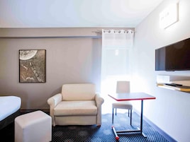 Habitación de hotel con sofá blanco, una mesita, televisor, un cuadro y una ventana con cortinas.