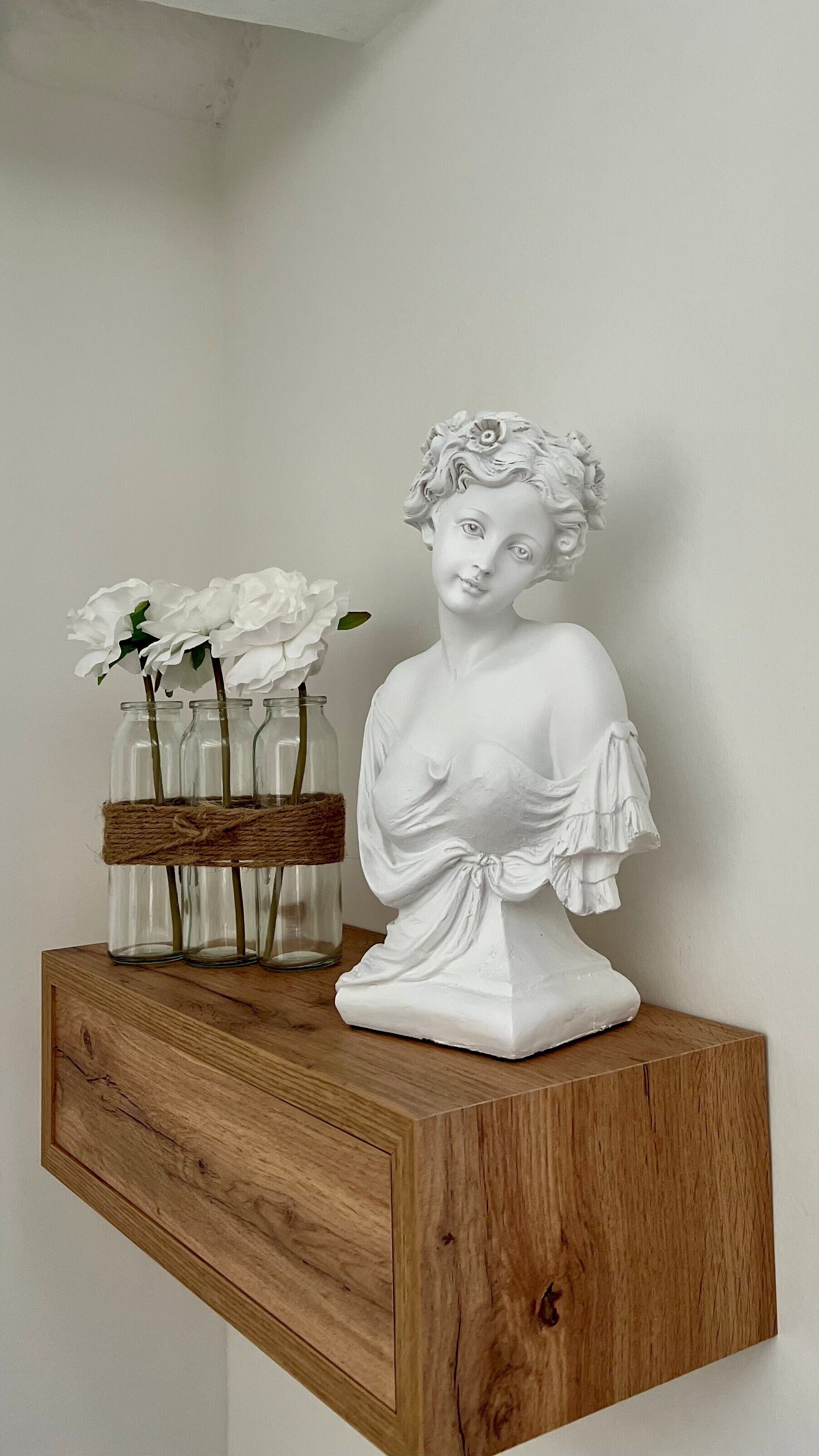 Un buste blanc, sculpté, est posé sur une étagère en bois, et un panier tressé, contenant des fleurs blanches, se trouve à proximité.