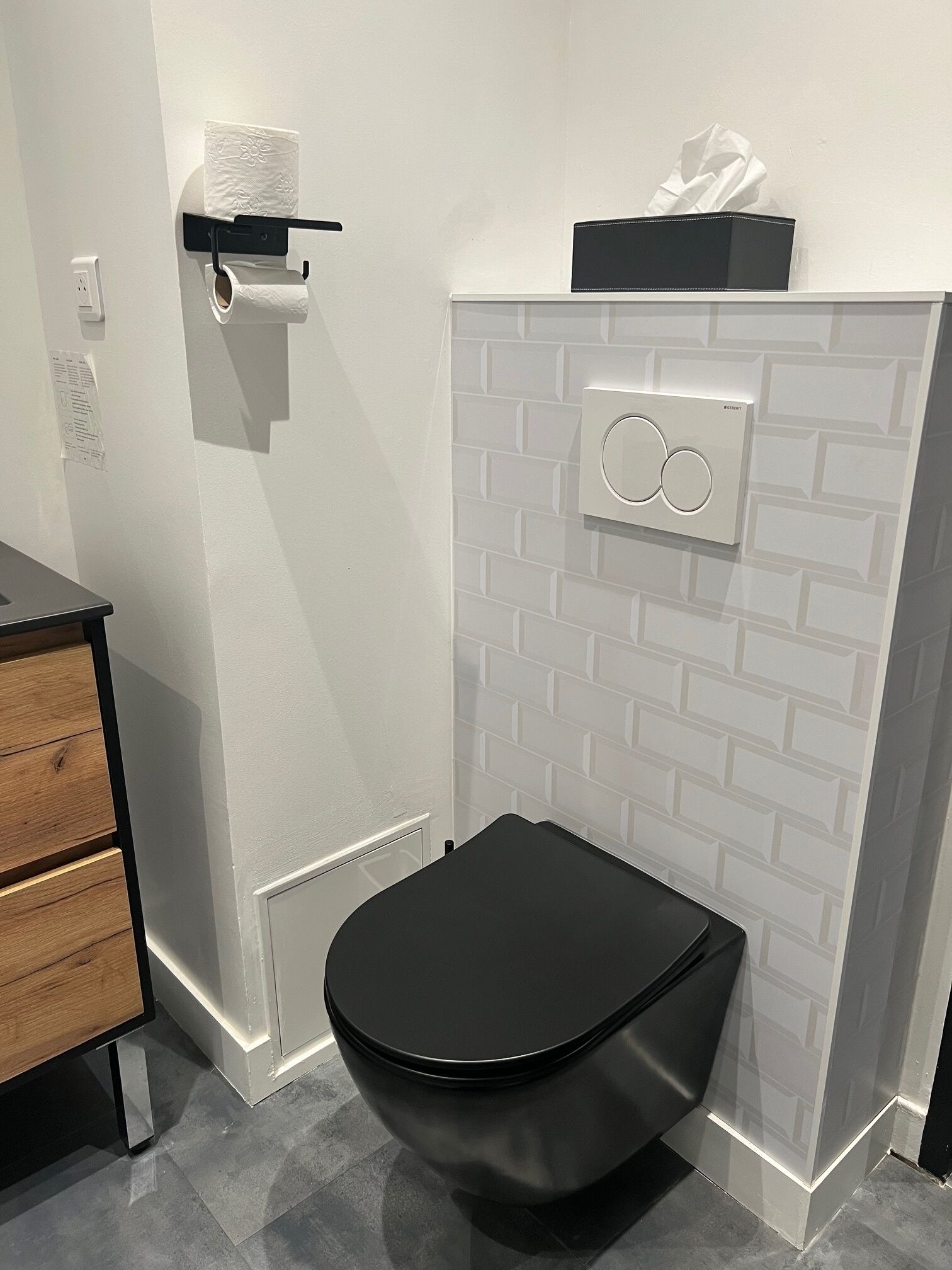 A modern bathroom with a black toilet, white tiled walls, and a black shelf with a tissue box.