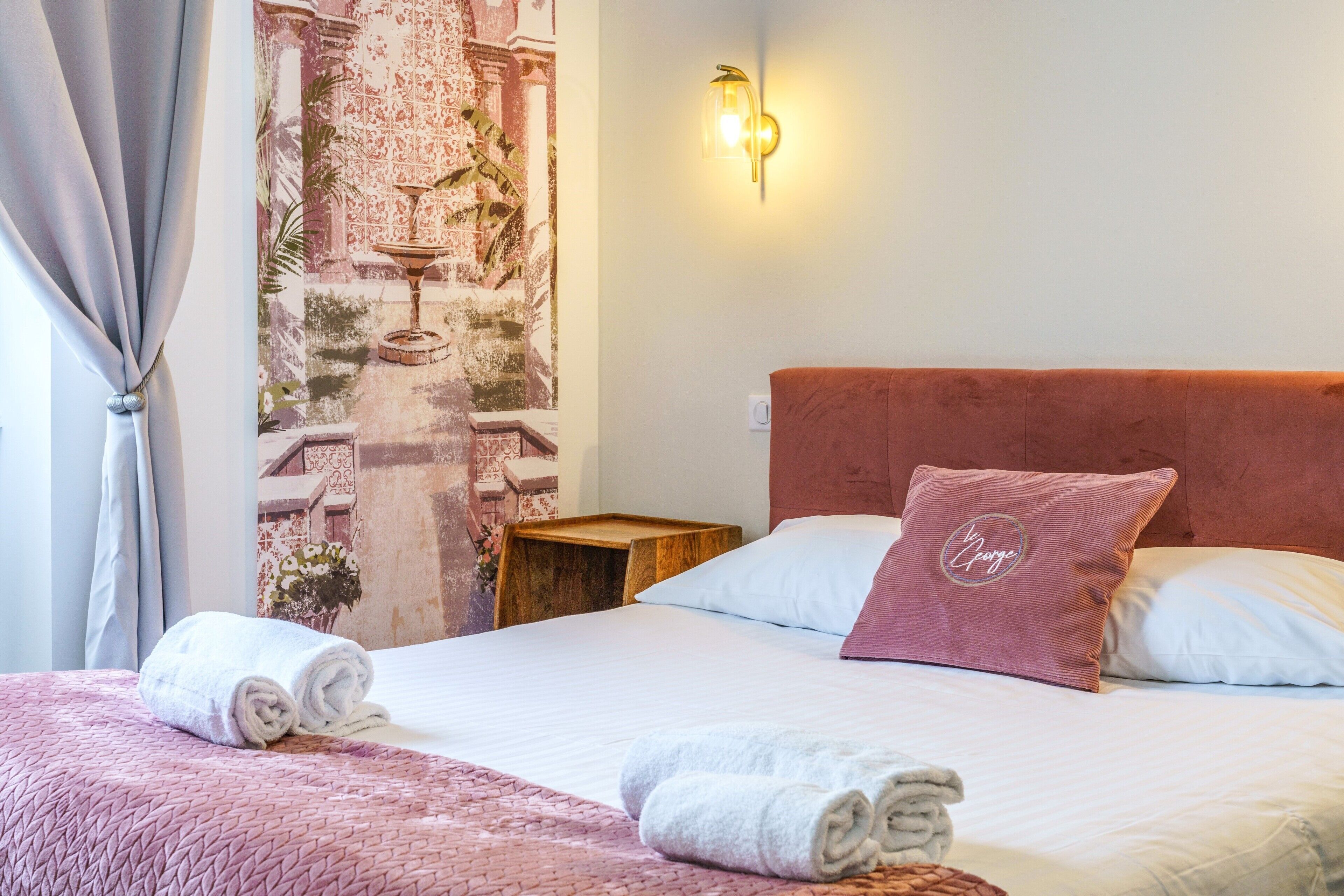Un lit bien fait, avec des serviettes pliées, une table de chevet en bois, une lampe fixée au mur et un coussin décoratif portant un nom.
